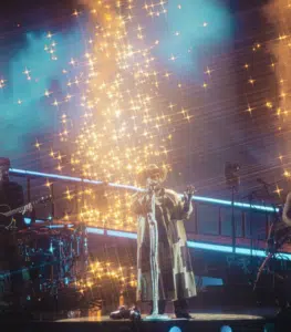 A band performs on stage under dramatic lighting effects, with golden sparks and beams of light illuminating the New York music scene.